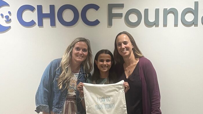 Esabella in front of the CHOC Foundation sign holding up one of her custom-created bags that reads "I can do hard things."